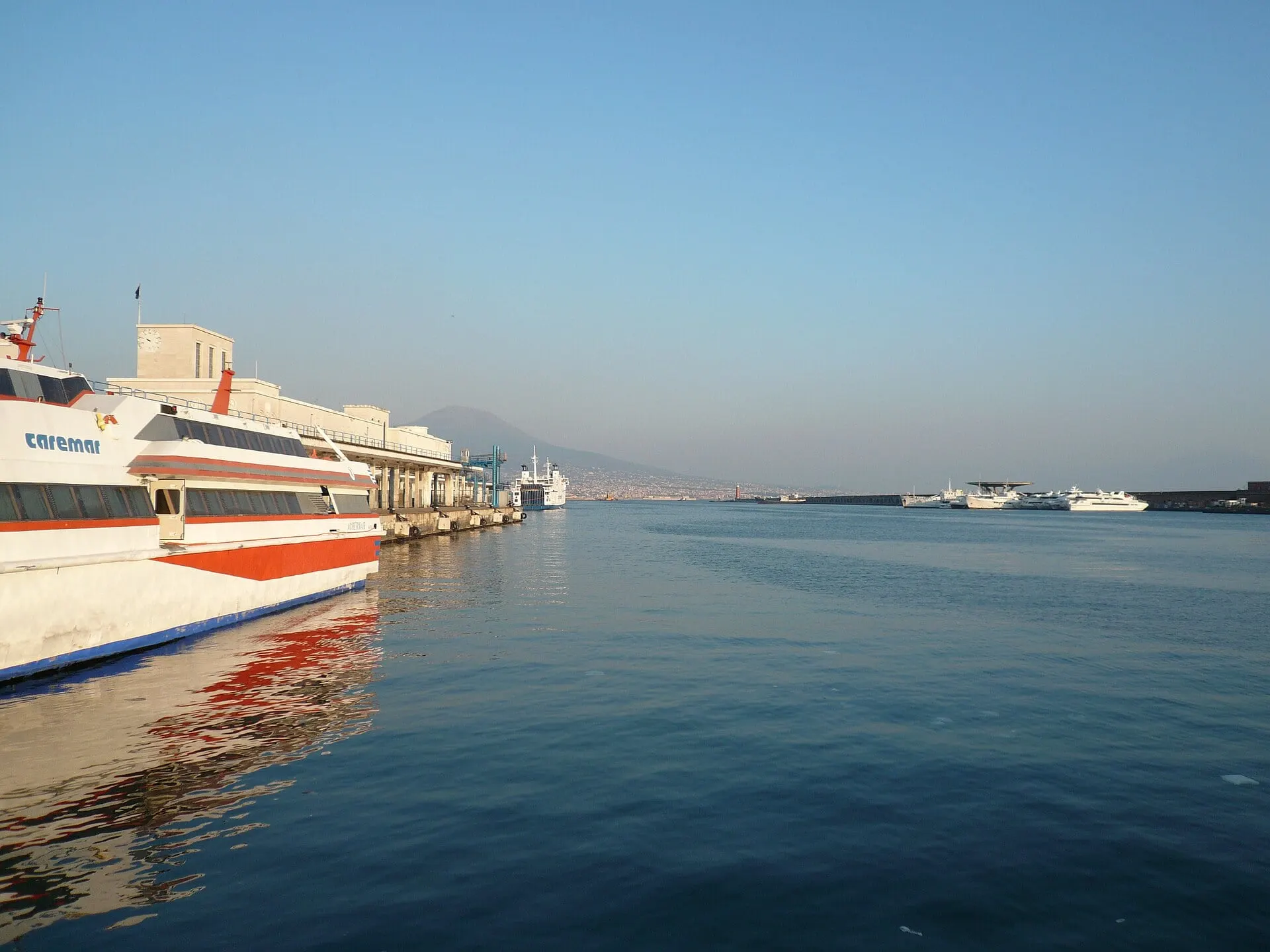 A Caremar fast boat moored at Molo Beverello in Naples with Vesuvius on the skyline across the harbour