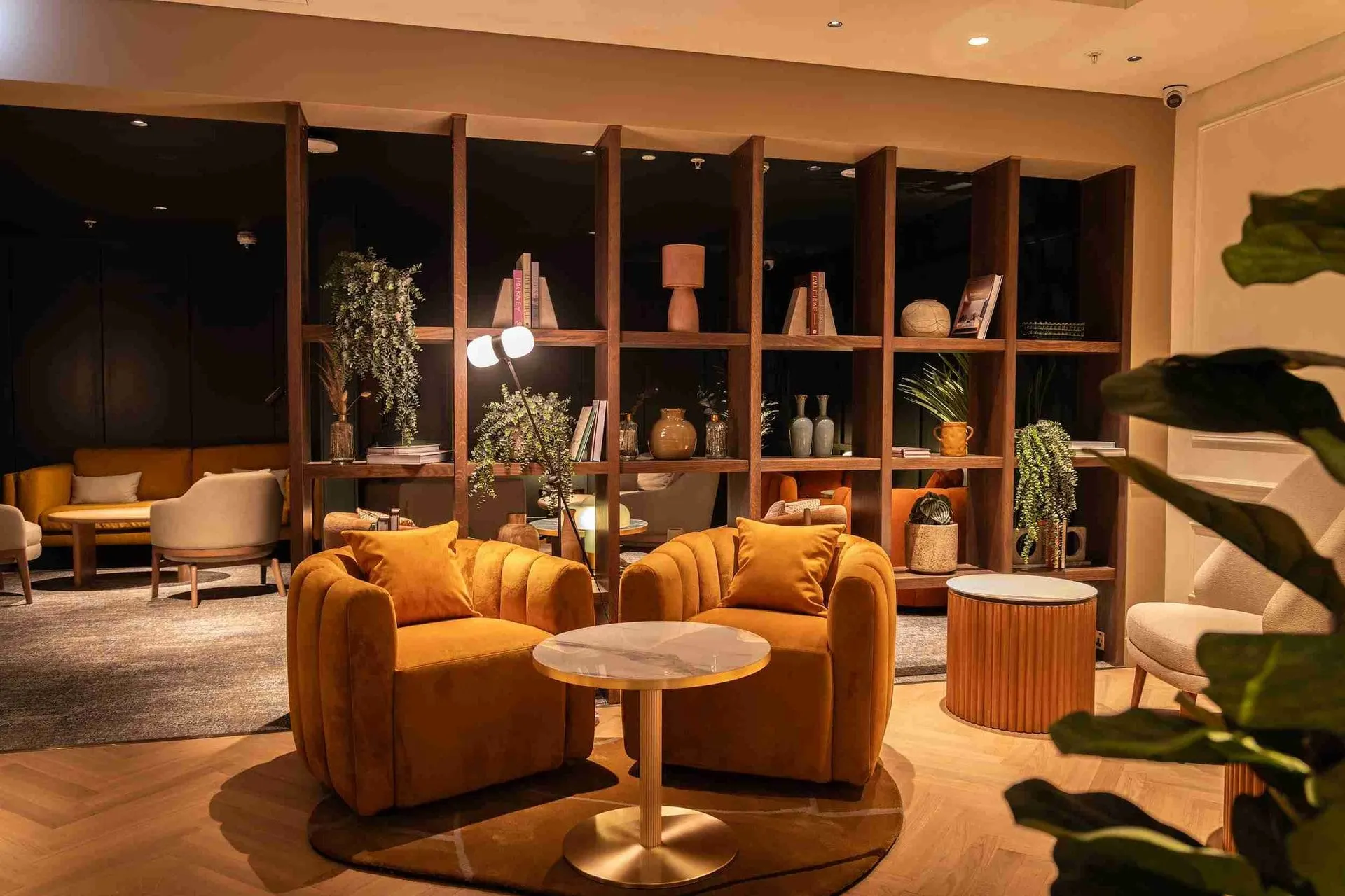 Two mustard velvet armchairs by a round marble table in front of a shelf of plants and ornaments