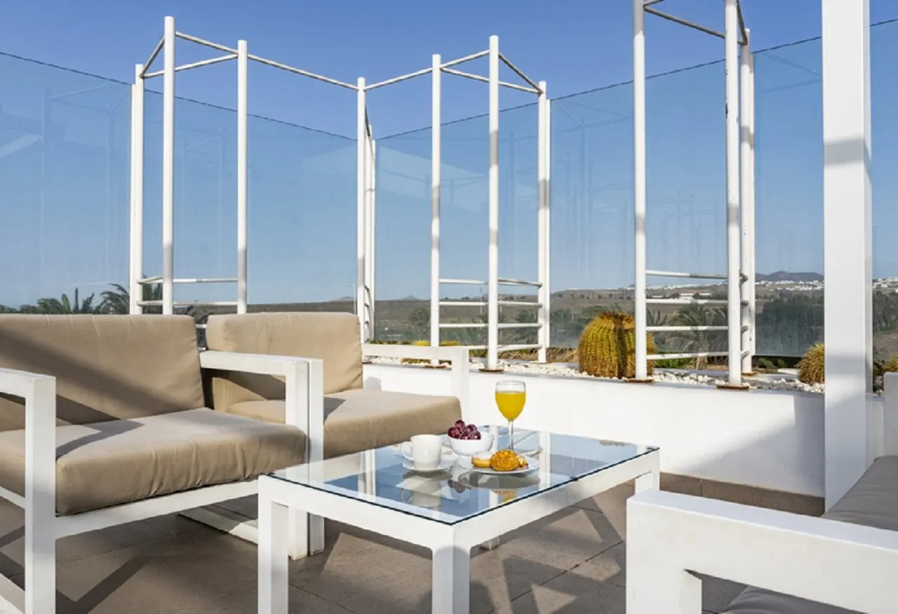 Glass table with orange juice, coffee and pastries beside terrace sofas and potted cacti in the sun