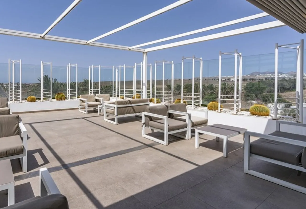 Grey cushioned sofas on a sunny roof terrace with glass screens, cacti and views over palms to the hills