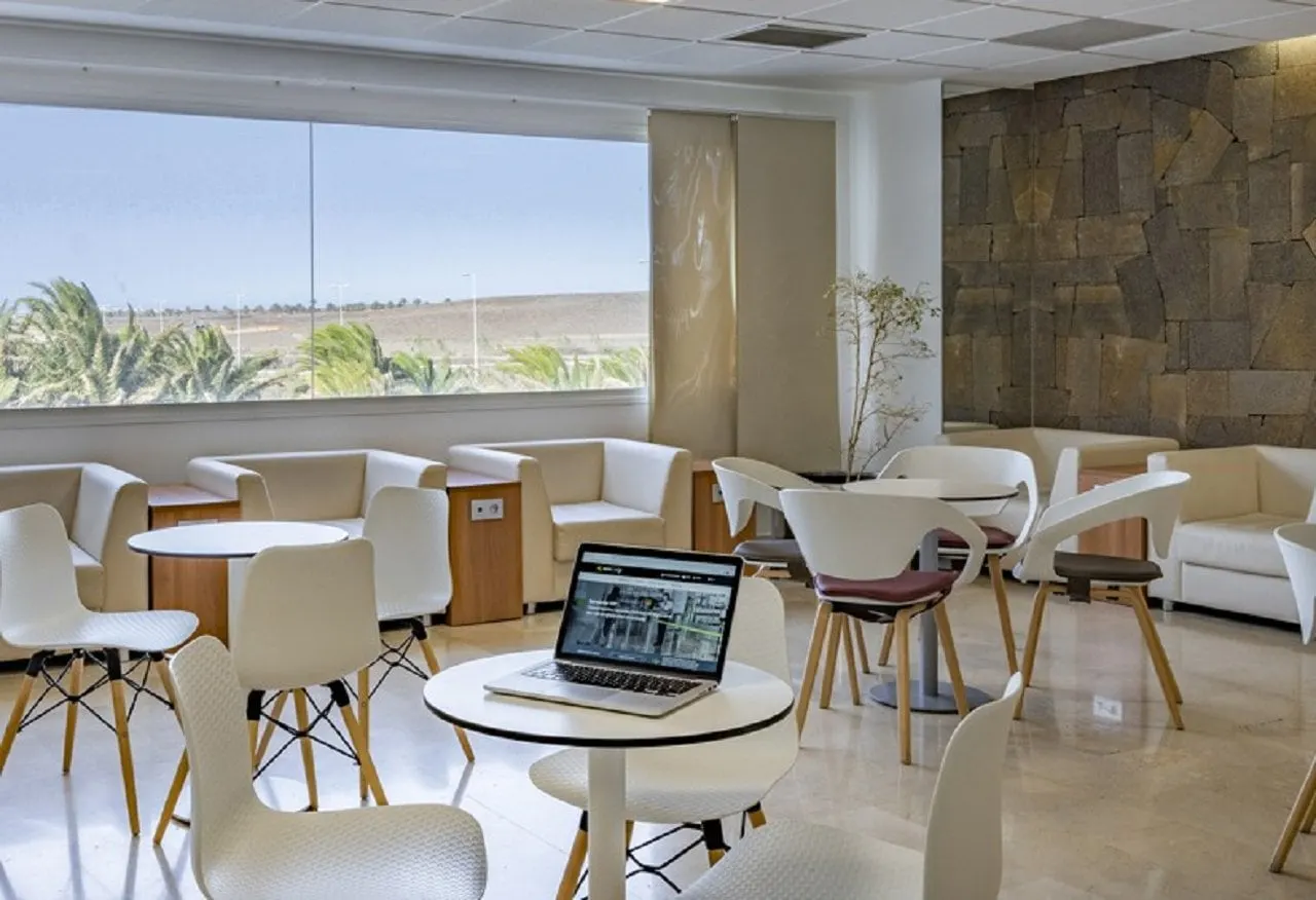 Laptop on a round table among white chairs and armchairs by a window over palms and a stone wall