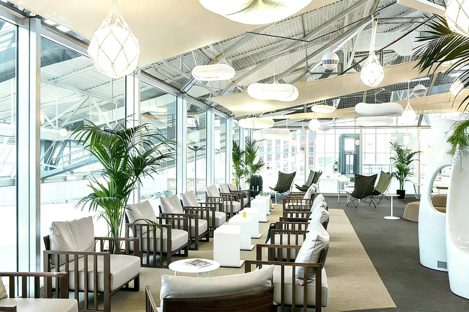 Bright lounge with white armchairs, potted palms and tall windows under a glass roof