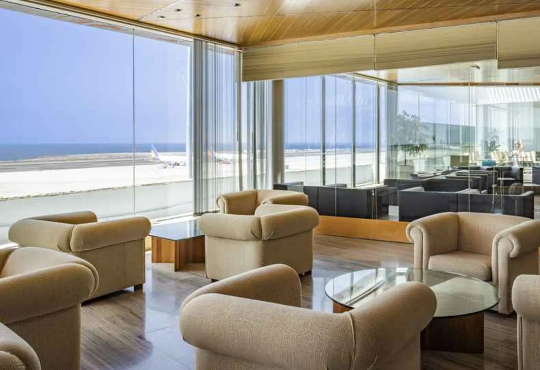 Beige armchairs round glass tables by floor-to-ceiling windows looking over the runway to the sea