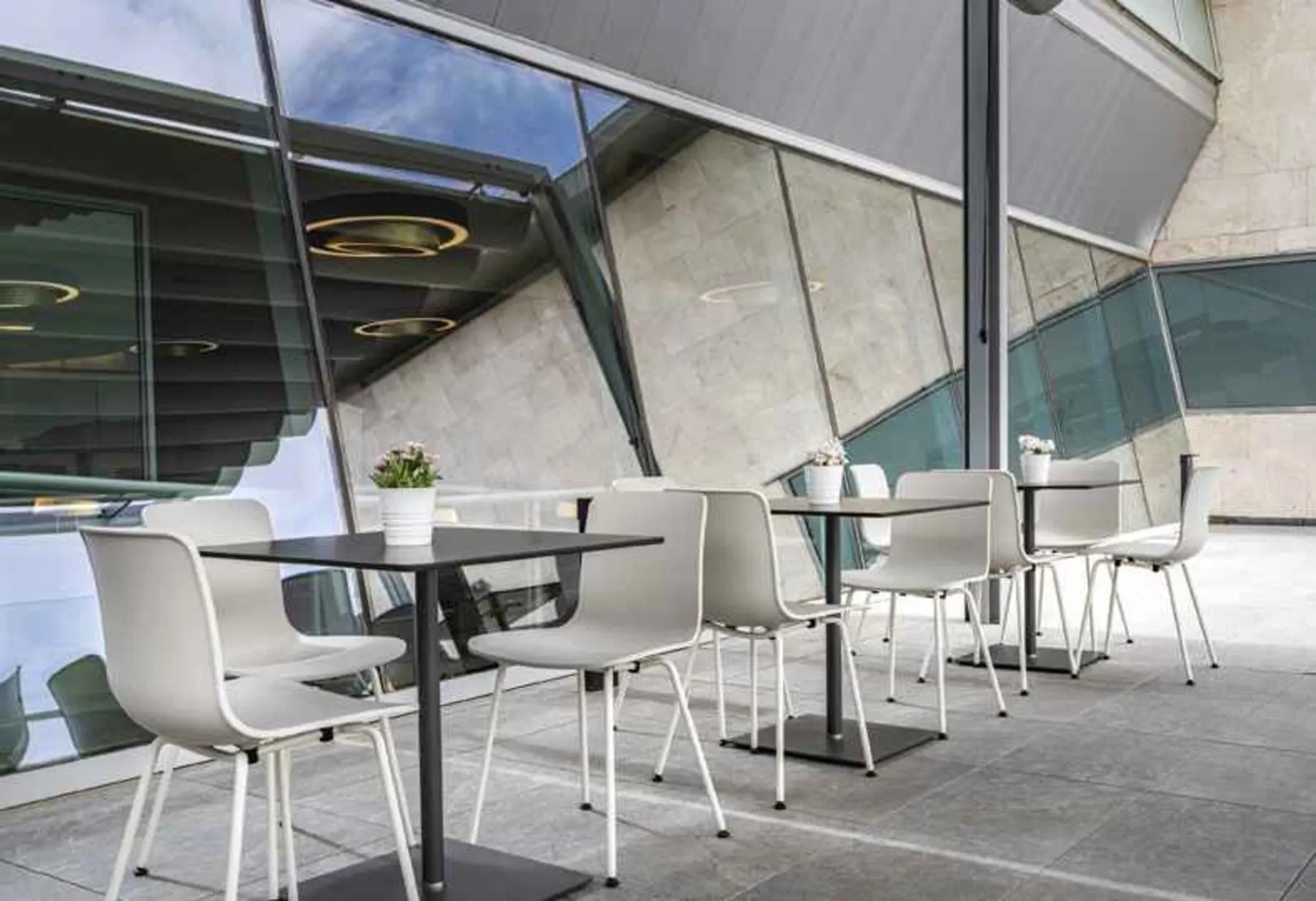 Chaises blanches et petites tables fleuries sur la terrasse extérieure, à côté de parois vitrées inclinées