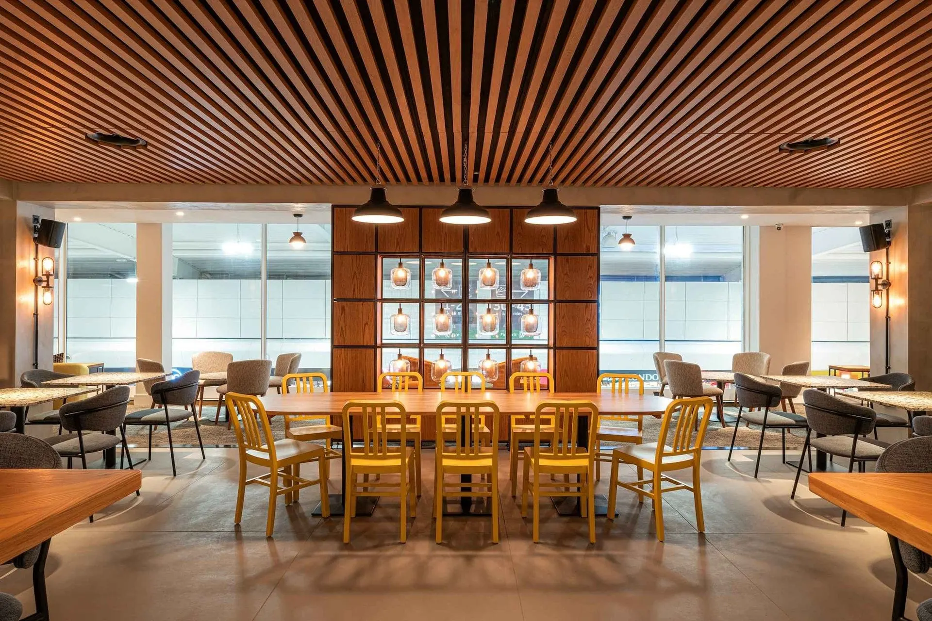 Long communal table with yellow chairs under pendant lamps and a slatted wooden ceiling