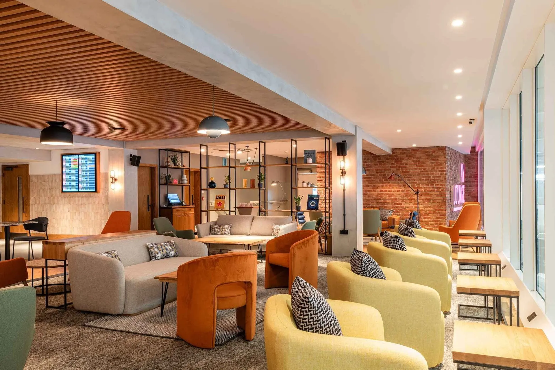 Cream sofas and yellow and orange armchairs under a slatted wooden ceiling, with shelves and a brick wall behind