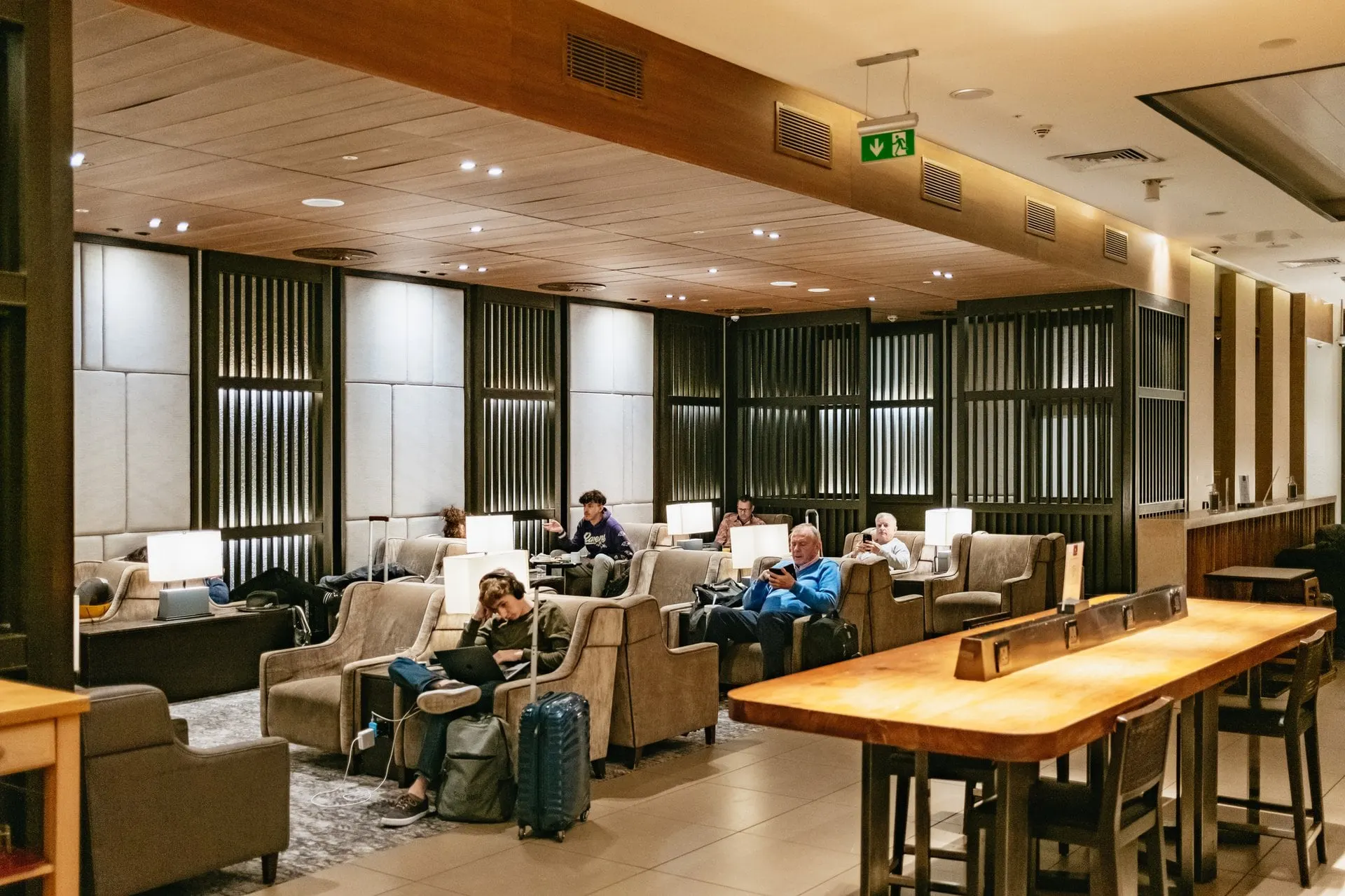 Guests in beige armchairs beside a long wooden table under a slatted wooden ceiling