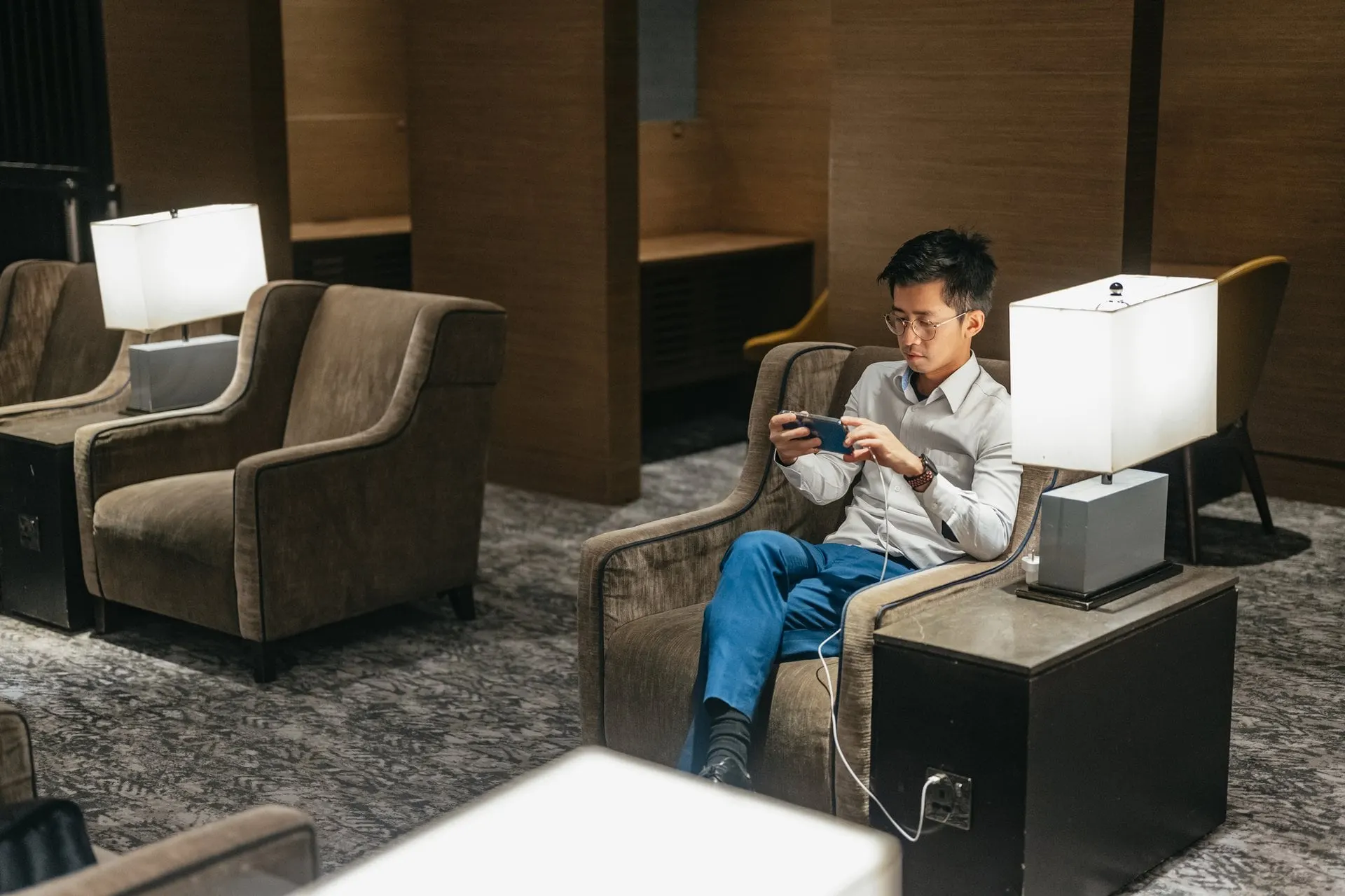 Man relaxing with his phone in a brown armchair beside a table lamp