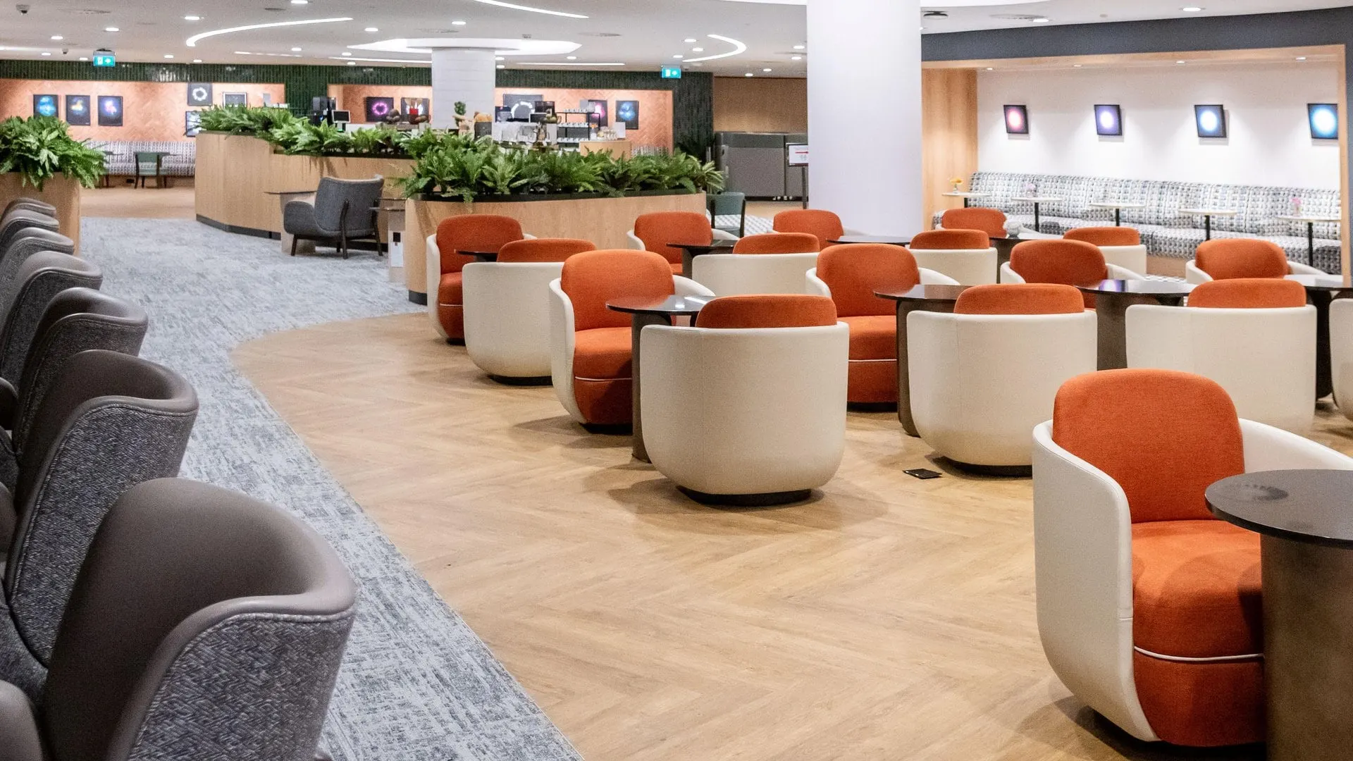Rows of white and orange tub chairs with low tables and planters in a wide, light Plaza Premium lounge