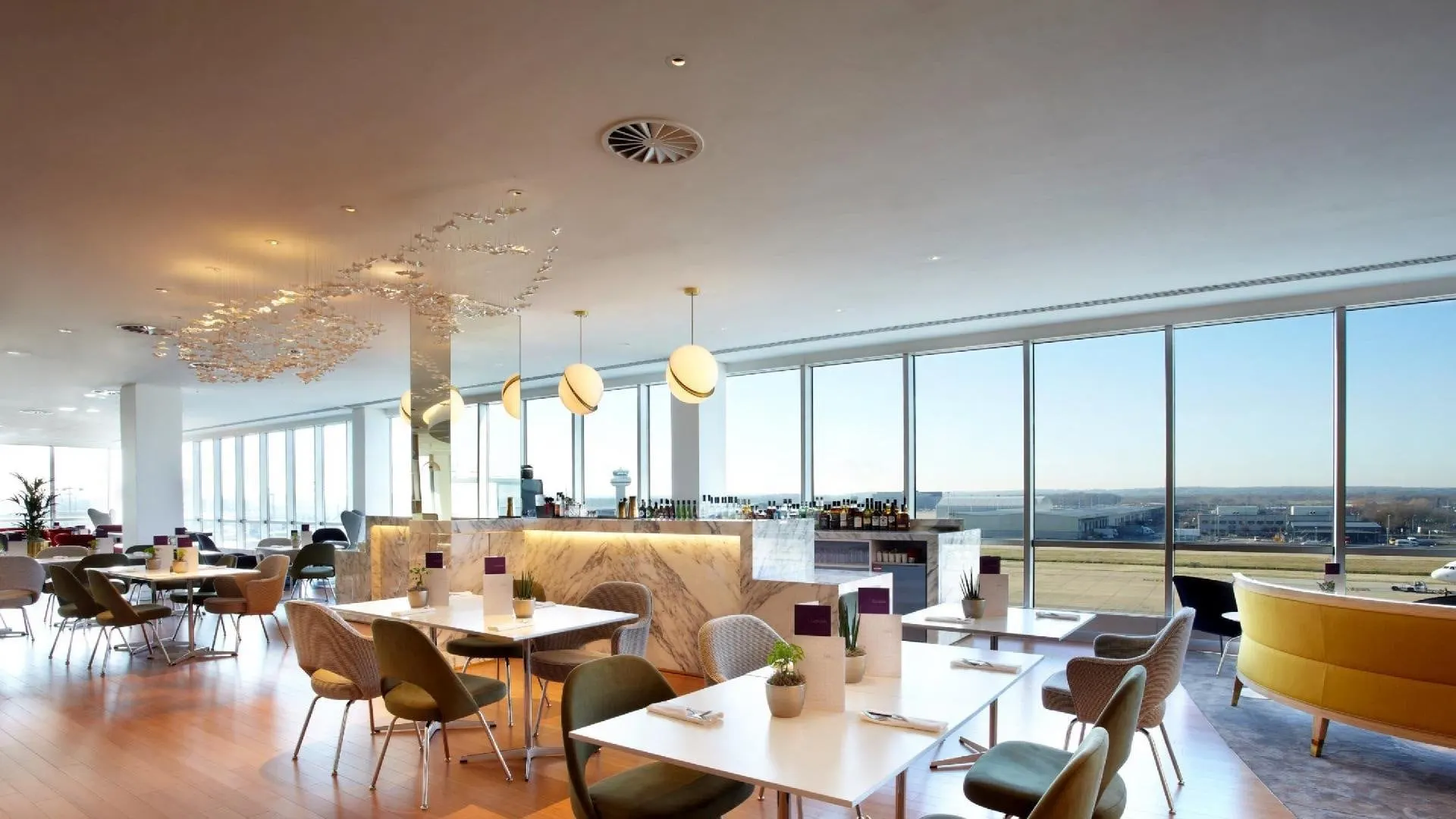 Dining tables and chairs by a wall of windows looking out over the airfield, with a bar on the left