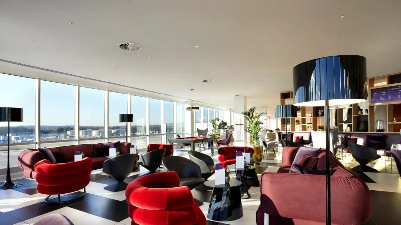 Red armchairs and sofas around low tables beside tall windows in a bright lounge