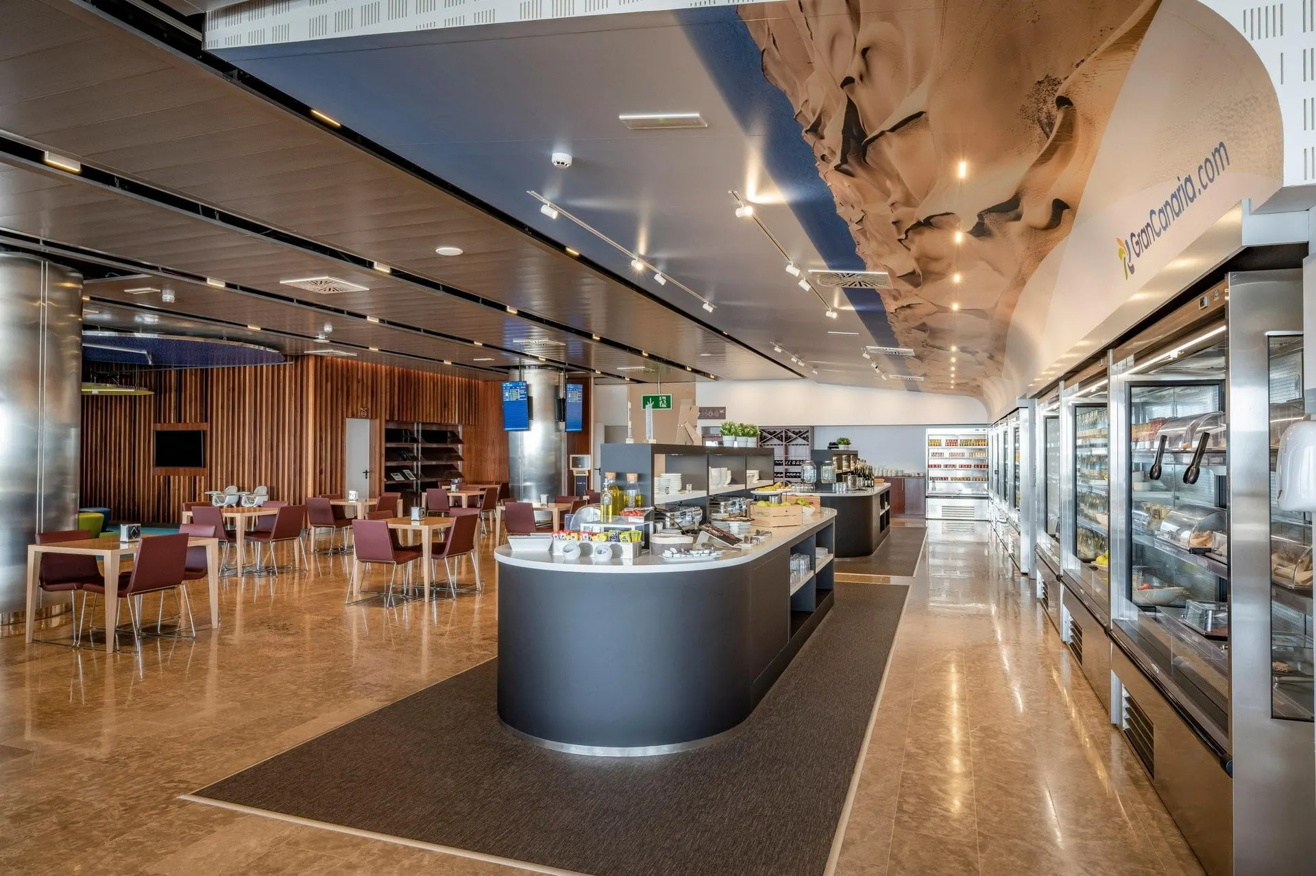 Curved buffet island and glass-fronted food counters beside dining tables under a wood ceiling