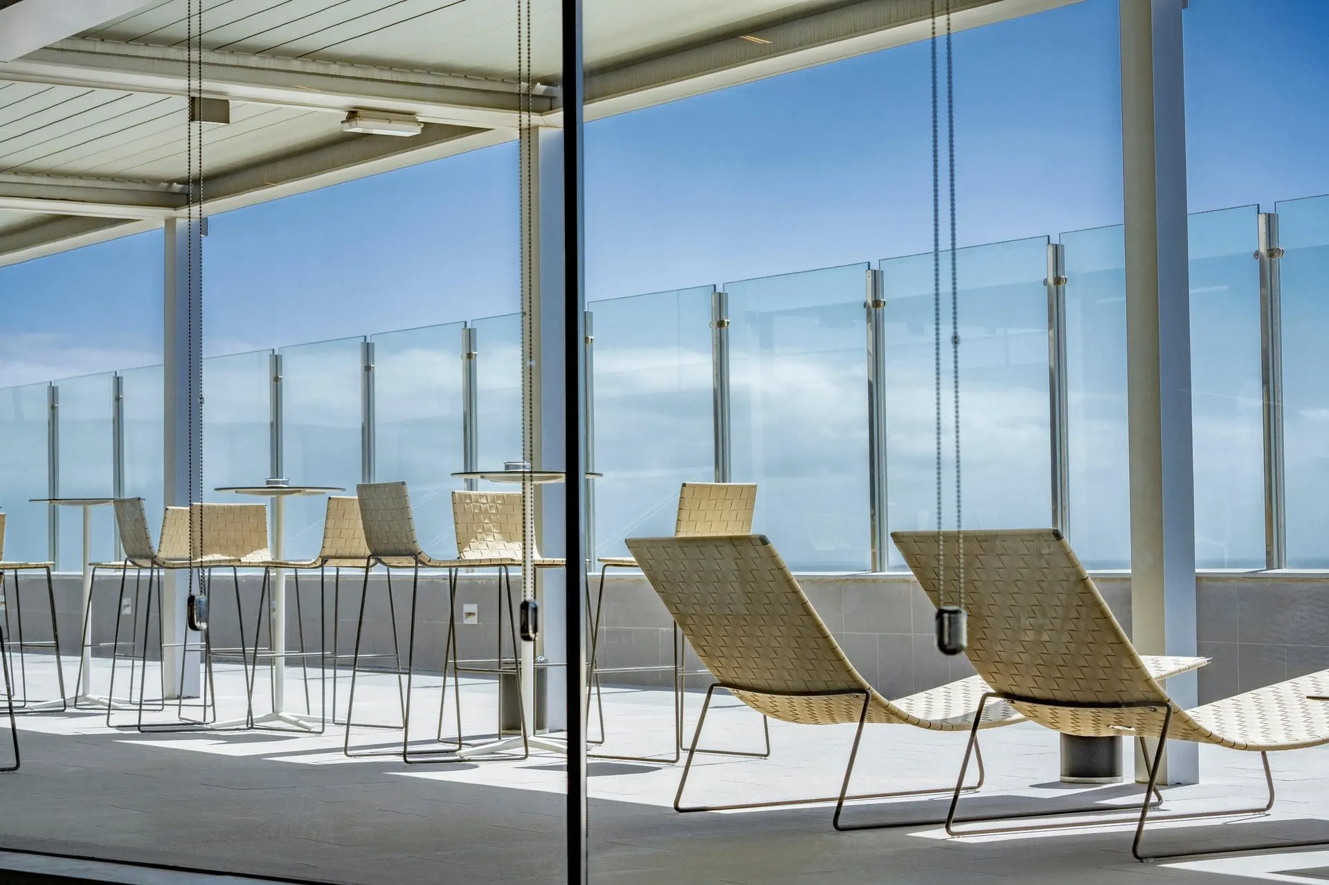 Woven loungers and high stools on the terrace behind a glass balustrade, with the sea and sky beyond