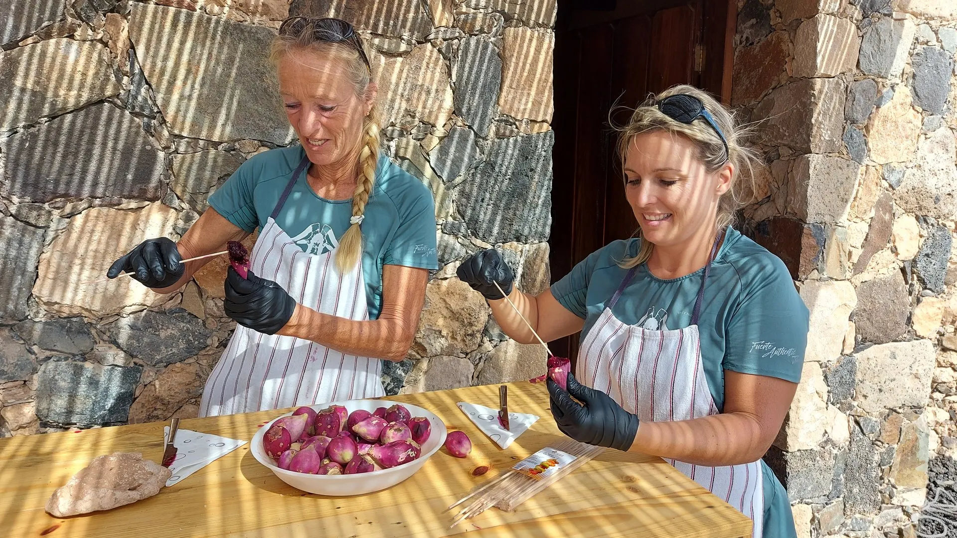 Two guides in aprons and gloves peeling prickly pears at a table against a stone wall