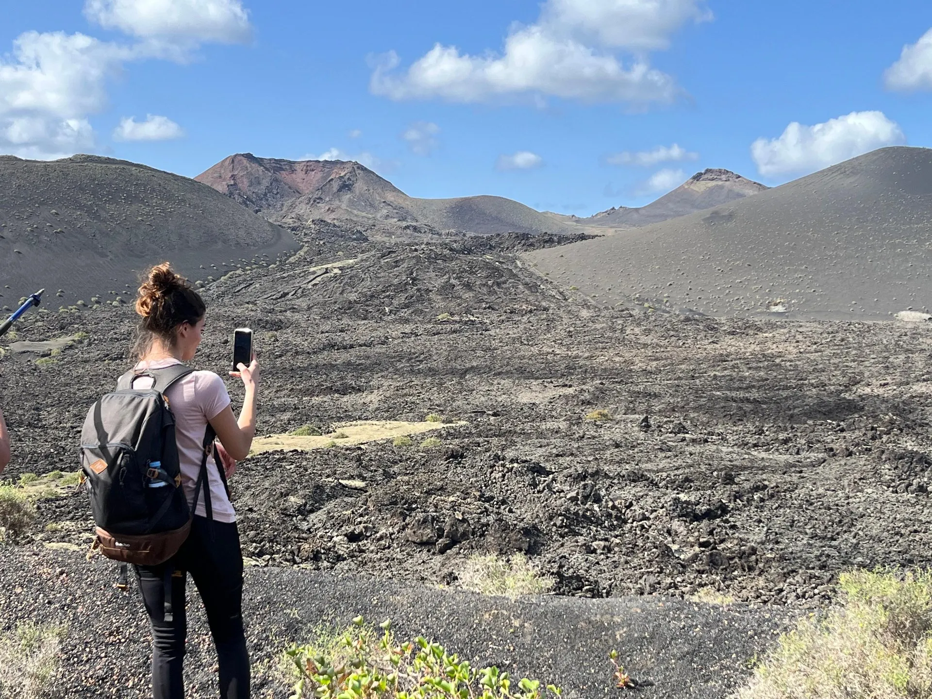 A hiker with a backpack photographs a field of black lava running up to red-brown volcanic cones
