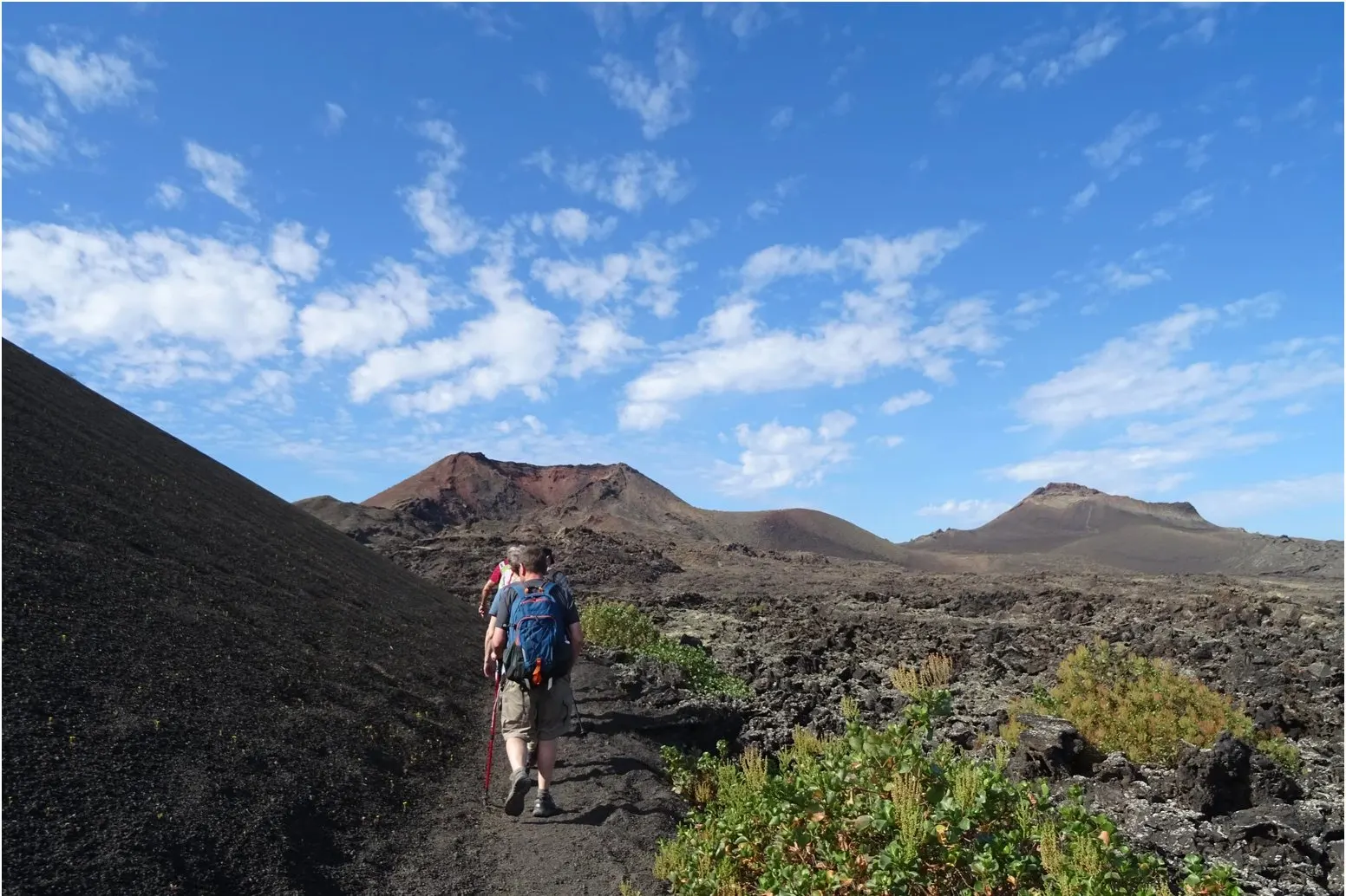 黒い火山の斜面沿いの火山灰の道を、赤い火口へ向かって歩く2人のハイカーと、溶岩原と緑の低木