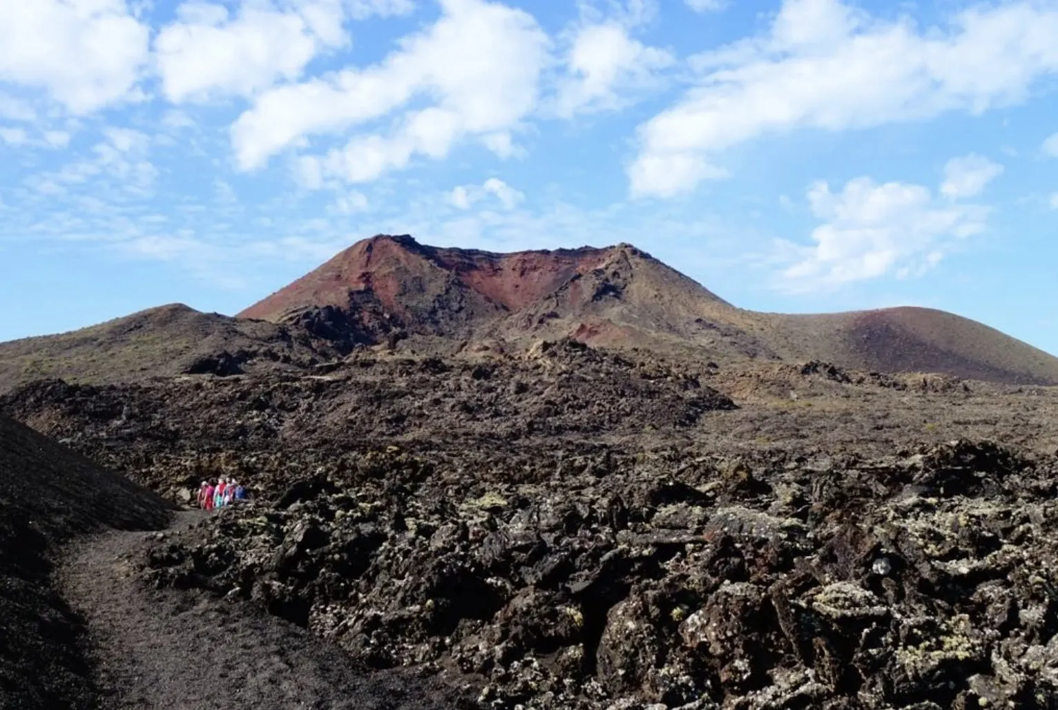 赤い壁面の火山錐の下、ごつごつした溶岩の中の道を歩く少人数のウォーカー