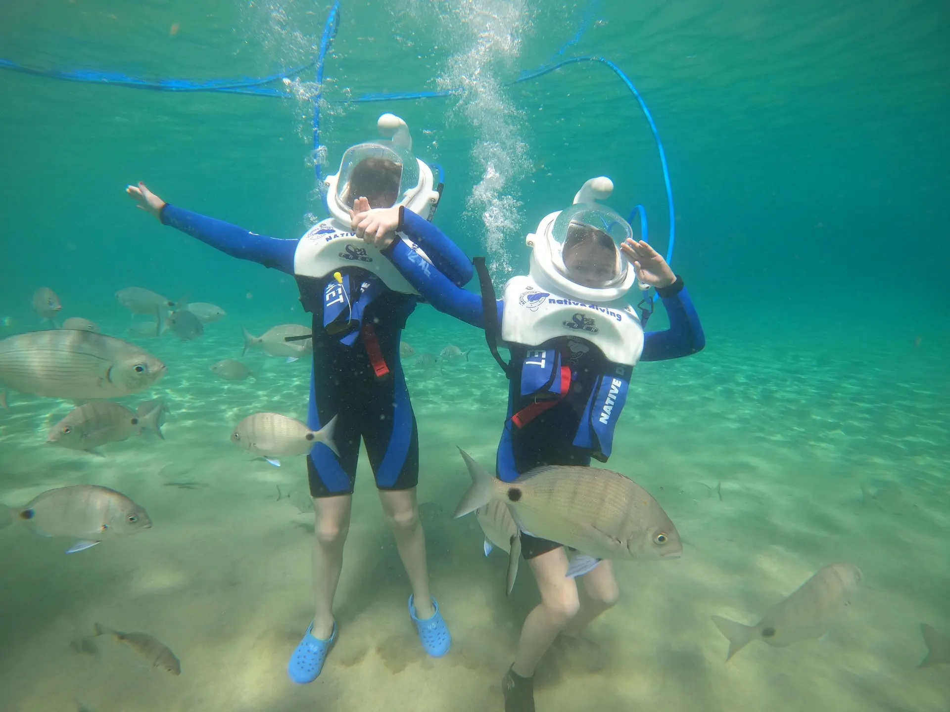 Two walkers in Sea Trek helmets waving on the sandy seabed