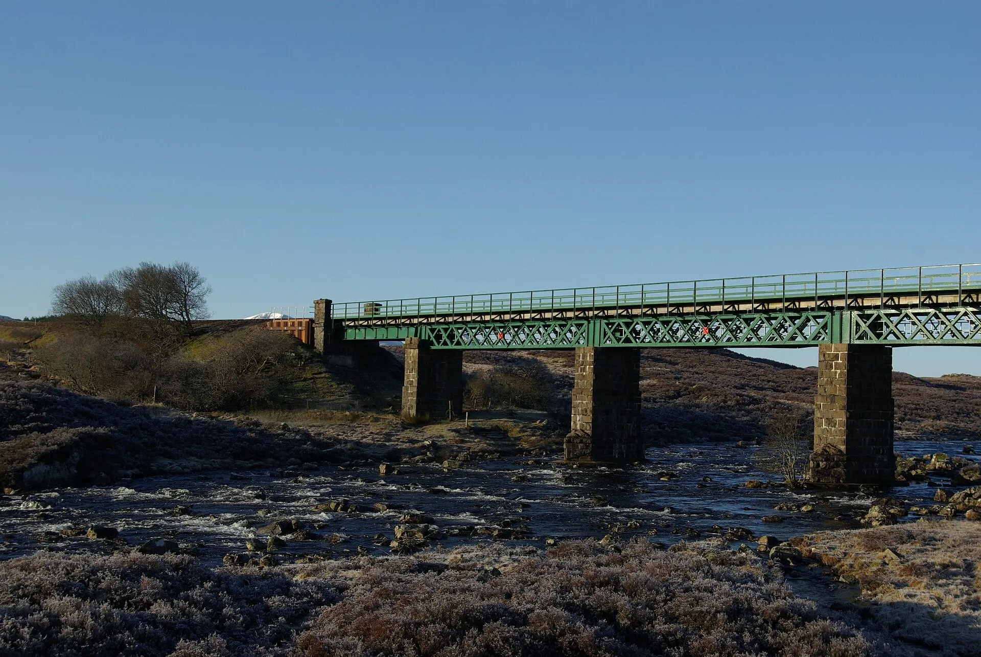 The green steel Gaur Viaduct carrying the West Highland Line across a river on Rannoch Moor under a clear blue sky.