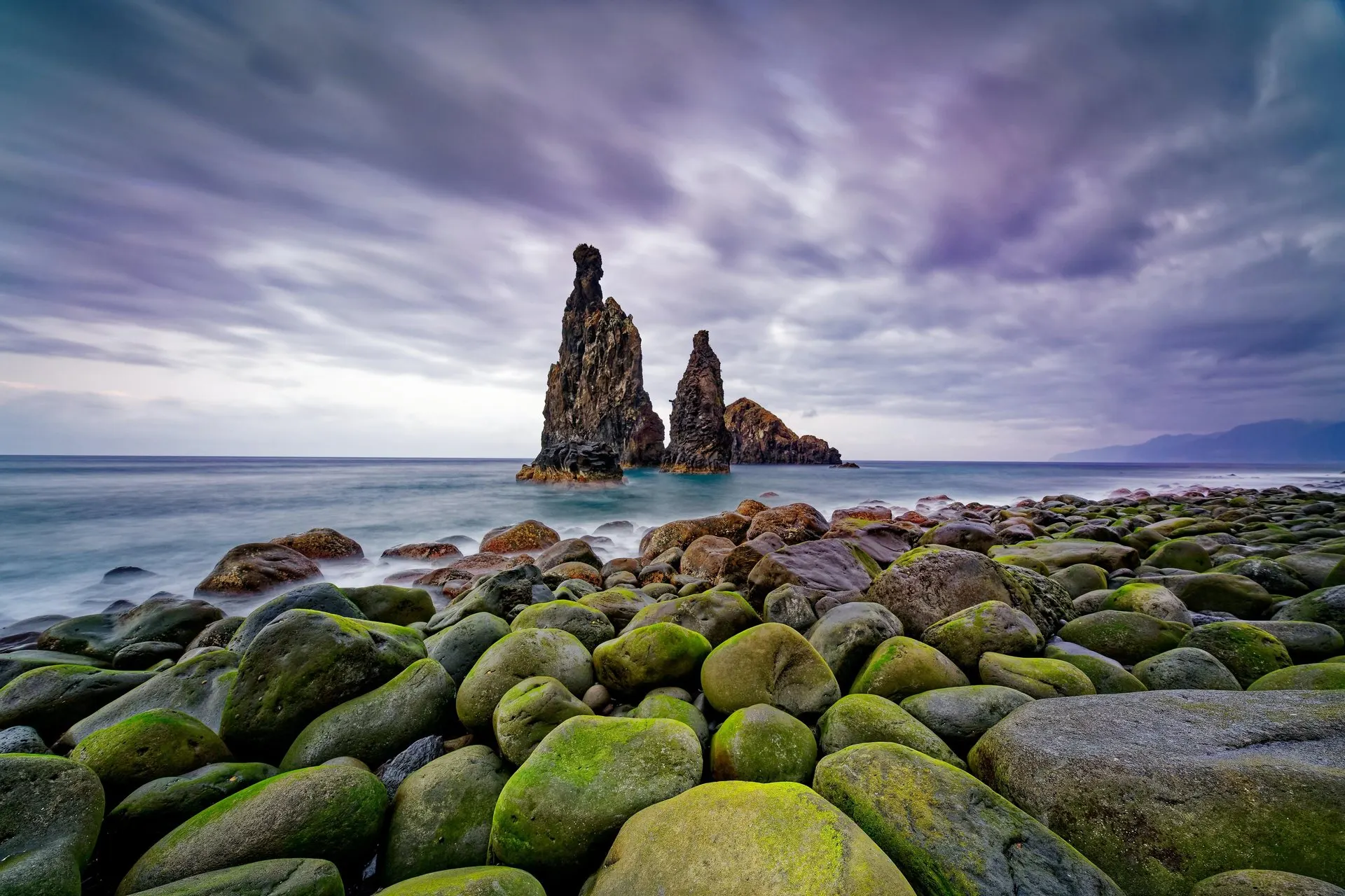 Basalt sea stacks off Ribeira da Janela, seen from a beach of large mossy boulders under a cloudy sky