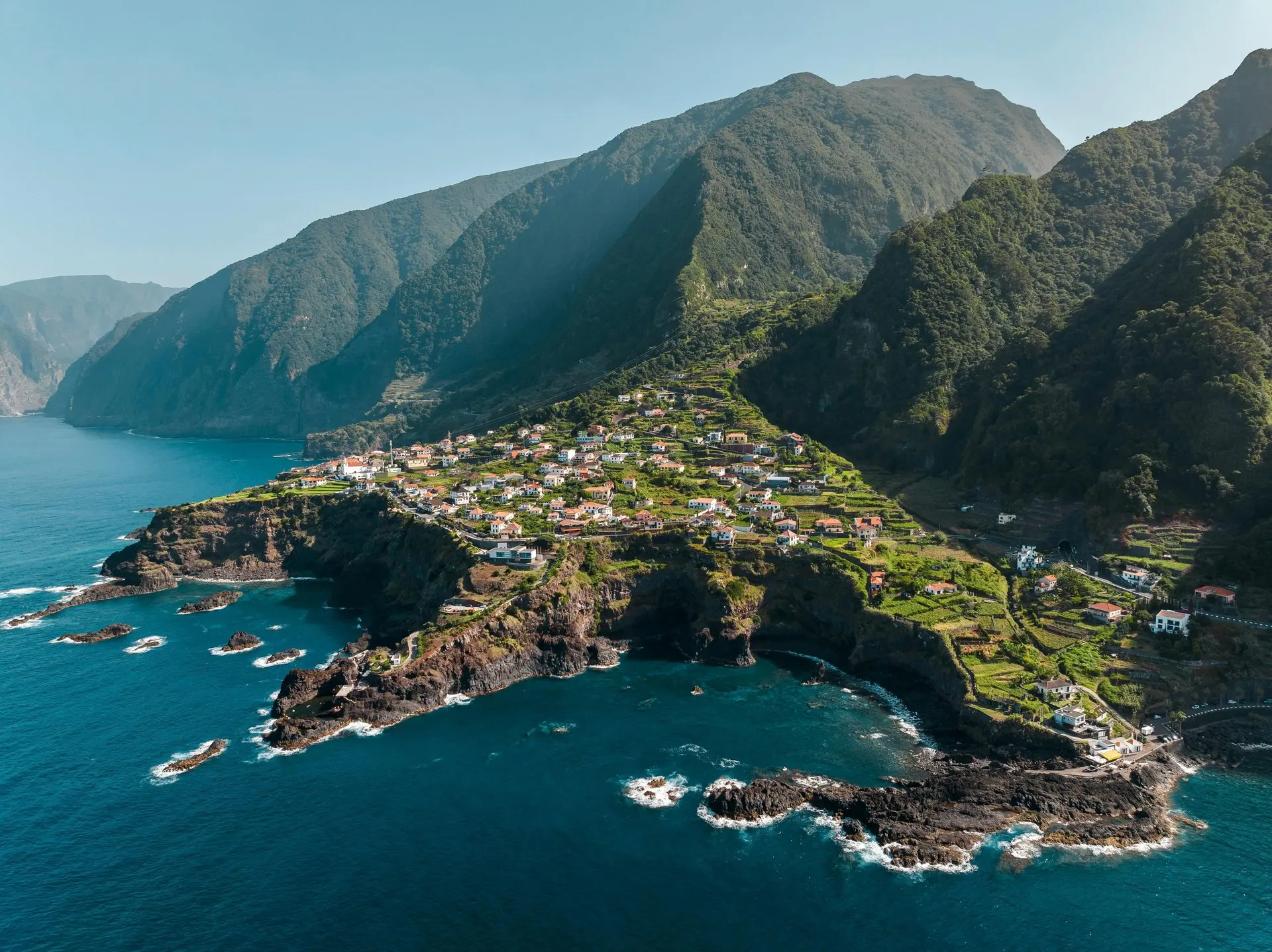 A village on a green headland on Madeira's north coast, below steep forested mountains, seen from the air
